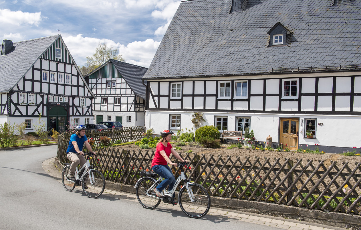 Zwei Radfahrer mit E-Bikes fahren durch ein Fachwerkdorf bei einer gef&uuml;hrten Radreise